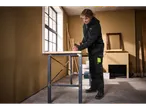 A man in a dark hoodie and work pants is sanding wood on a workbench in a room with a large window.