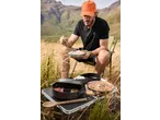 Man camping, eating from a plate, with camping cookware and food on a portable table.