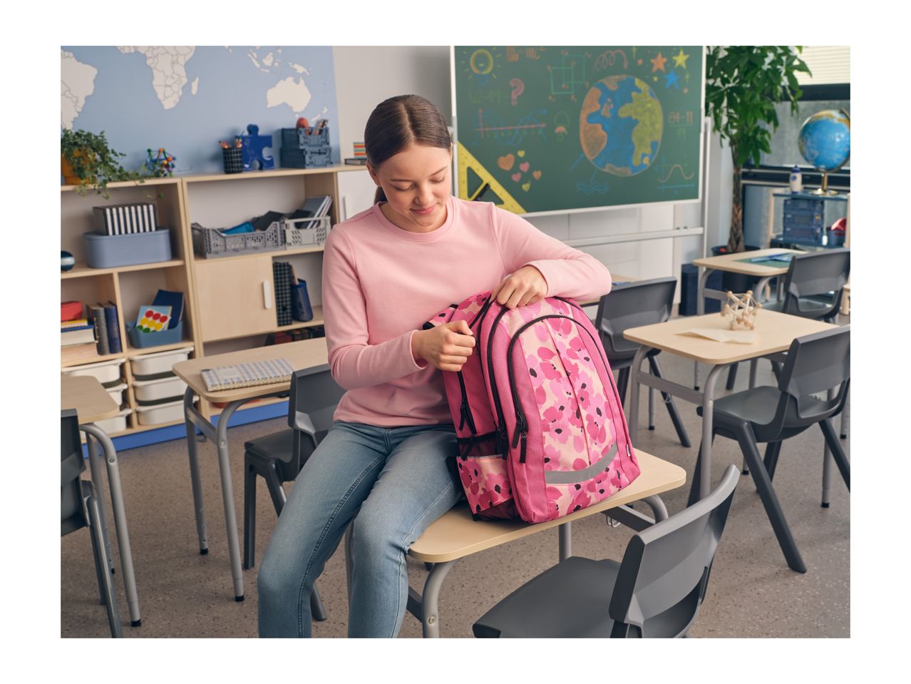A girl in a classroom, sitting on a desk and opening her pink floral backpack.