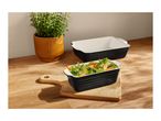 Black and white rectangular baking dishes, one filled with vegetables, on a wooden board.