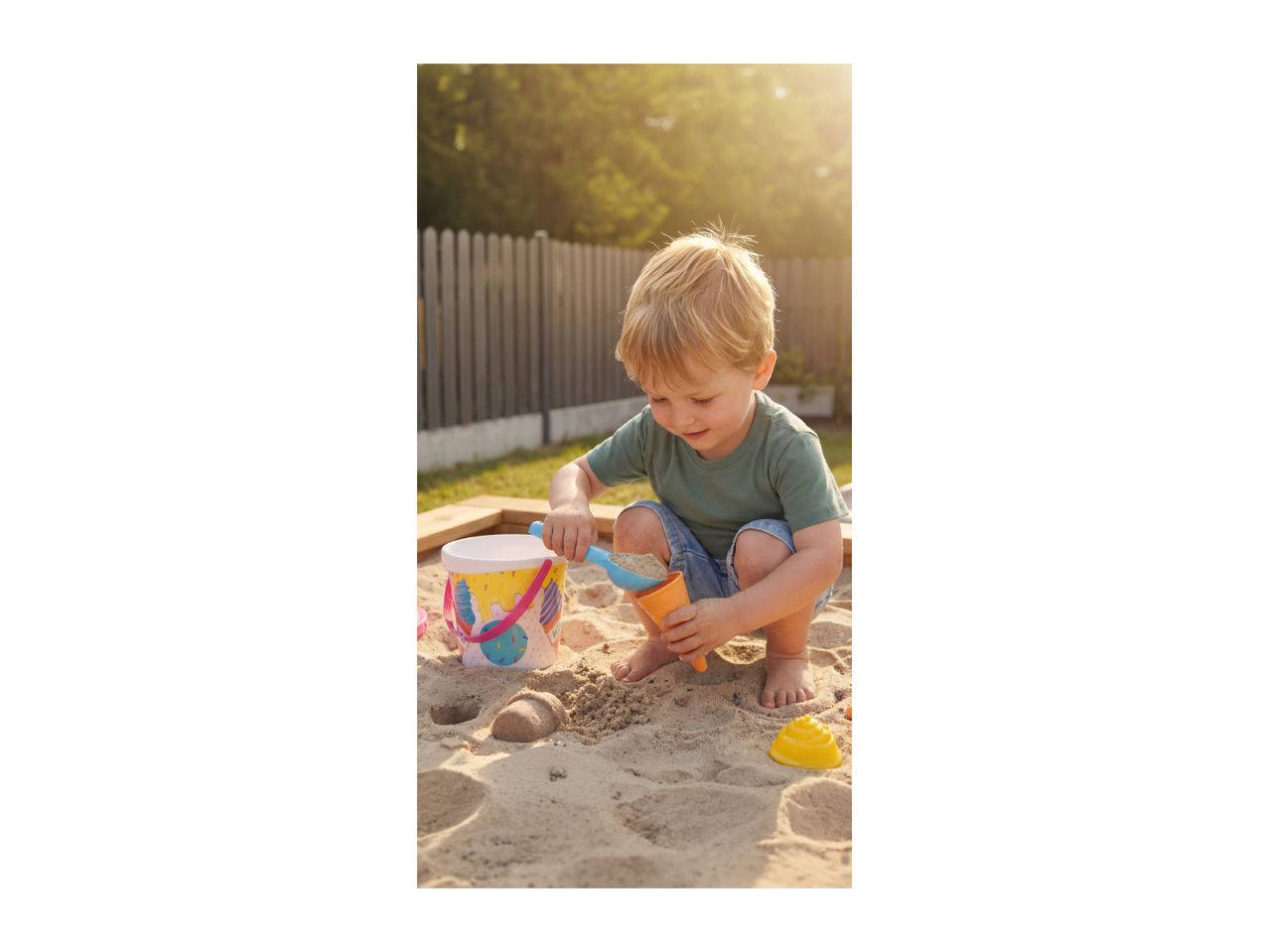 Toddler playing in a sandbox with a bucket, spade, and sand molds.