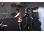 A laser level on a tripod projects green lines onto a wall where a man is marking measurements.