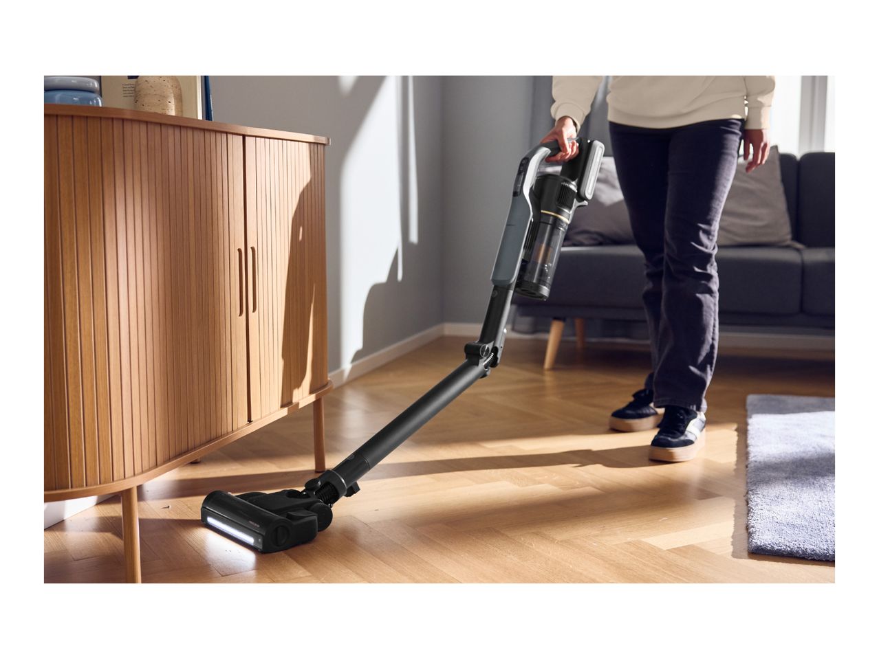 A person vacuuming a wooden floor under a cabinet with a cordless stick vacuum cleaner.