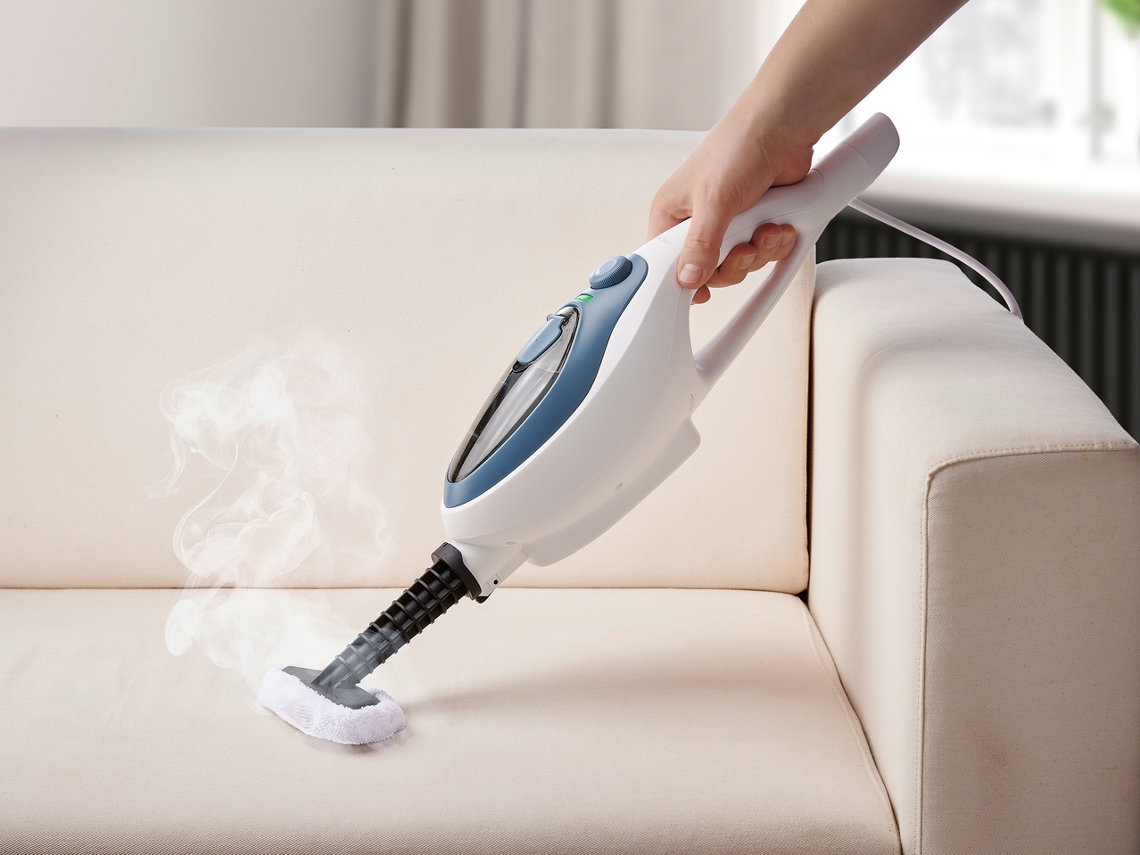 Πλυντήριο ατμού χειρός καθαρίζει έναν μπεζ καναπέ, με ορατό ατμό.