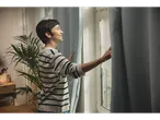 Femeie zâmbind, deschizând perdelele LIVARNO home pentru a lăsa lumina să intre.