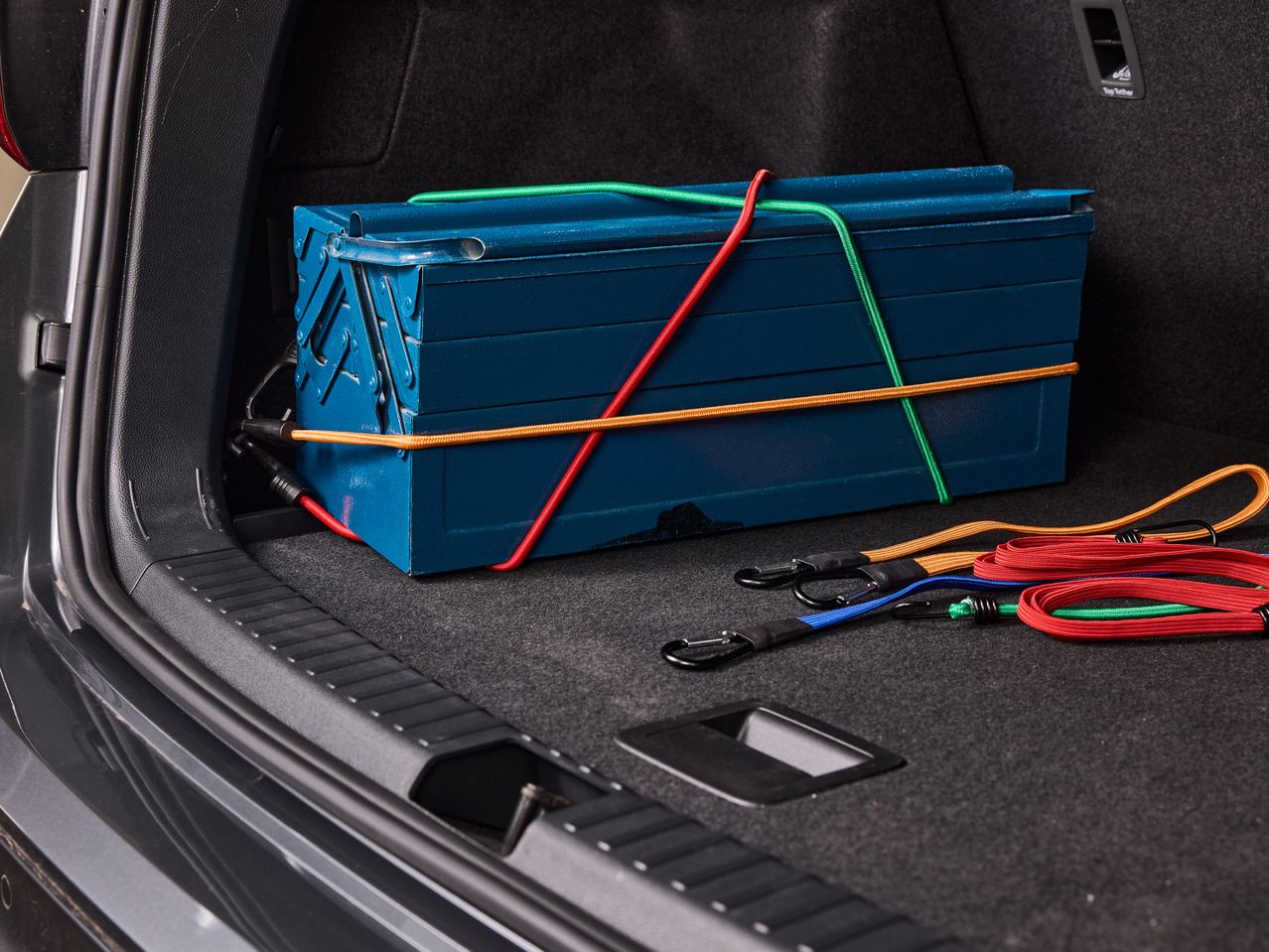 Cassetta degli attrezzi blu fissata con corde elastiche colorate nel bagagliaio di un'auto.
