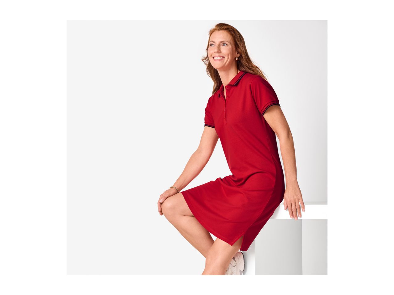 Woman in a red polo dress with striped cuffs and collar, smiling and looking up.