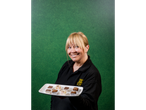 A woman in a black polo shirt holds a tray of brownies and other small desserts.