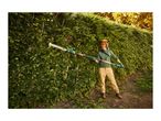 Person trimming a tall hedge with a long-reach hedge trimmer, wearing protective gear.