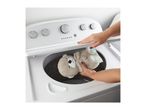 A person placing a grey teddy bear plush toy into a white top-loading washing machine.