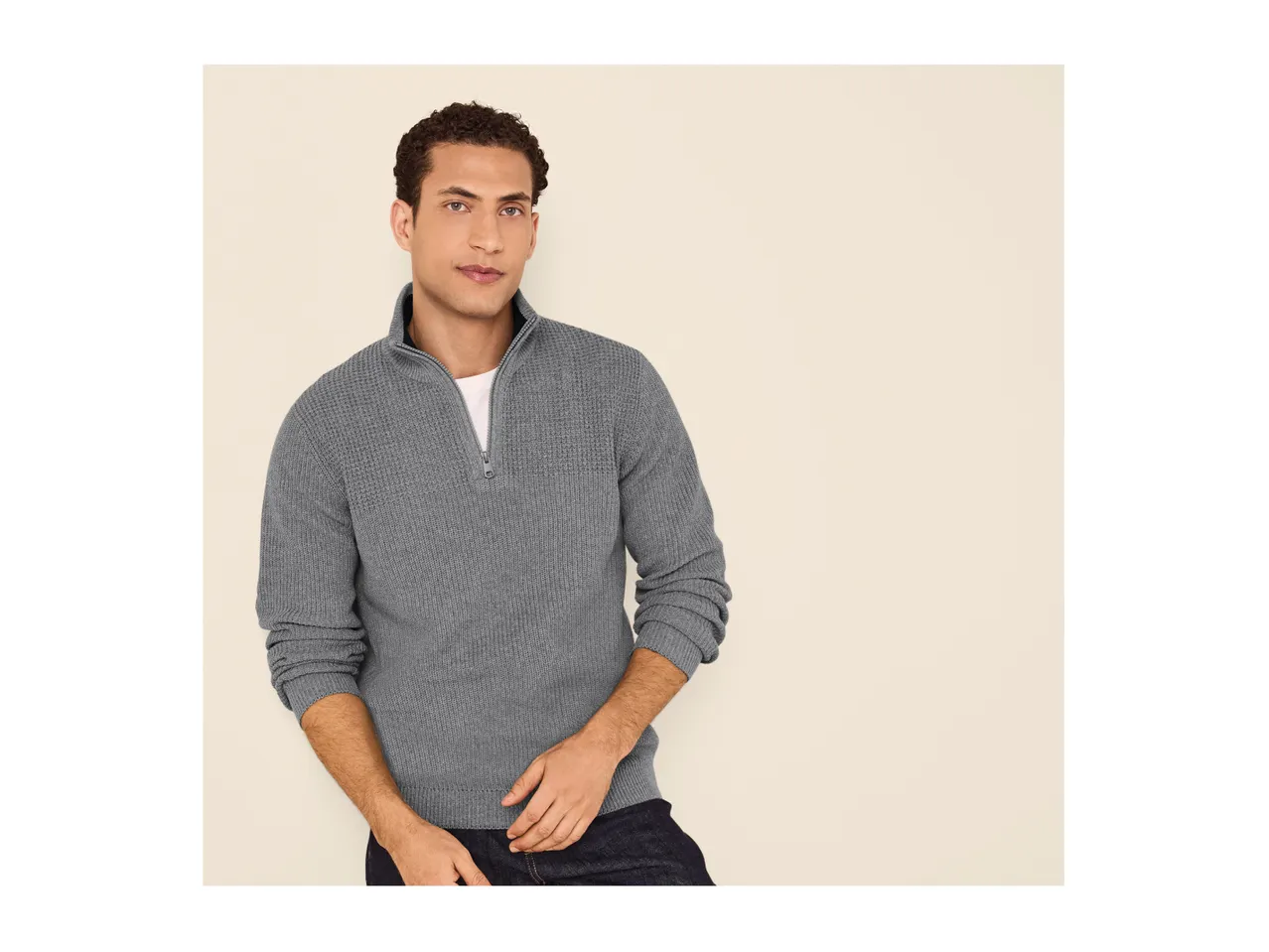 A man wearing a grey half-zip knitted jumper over a white t-shirt and dark jeans