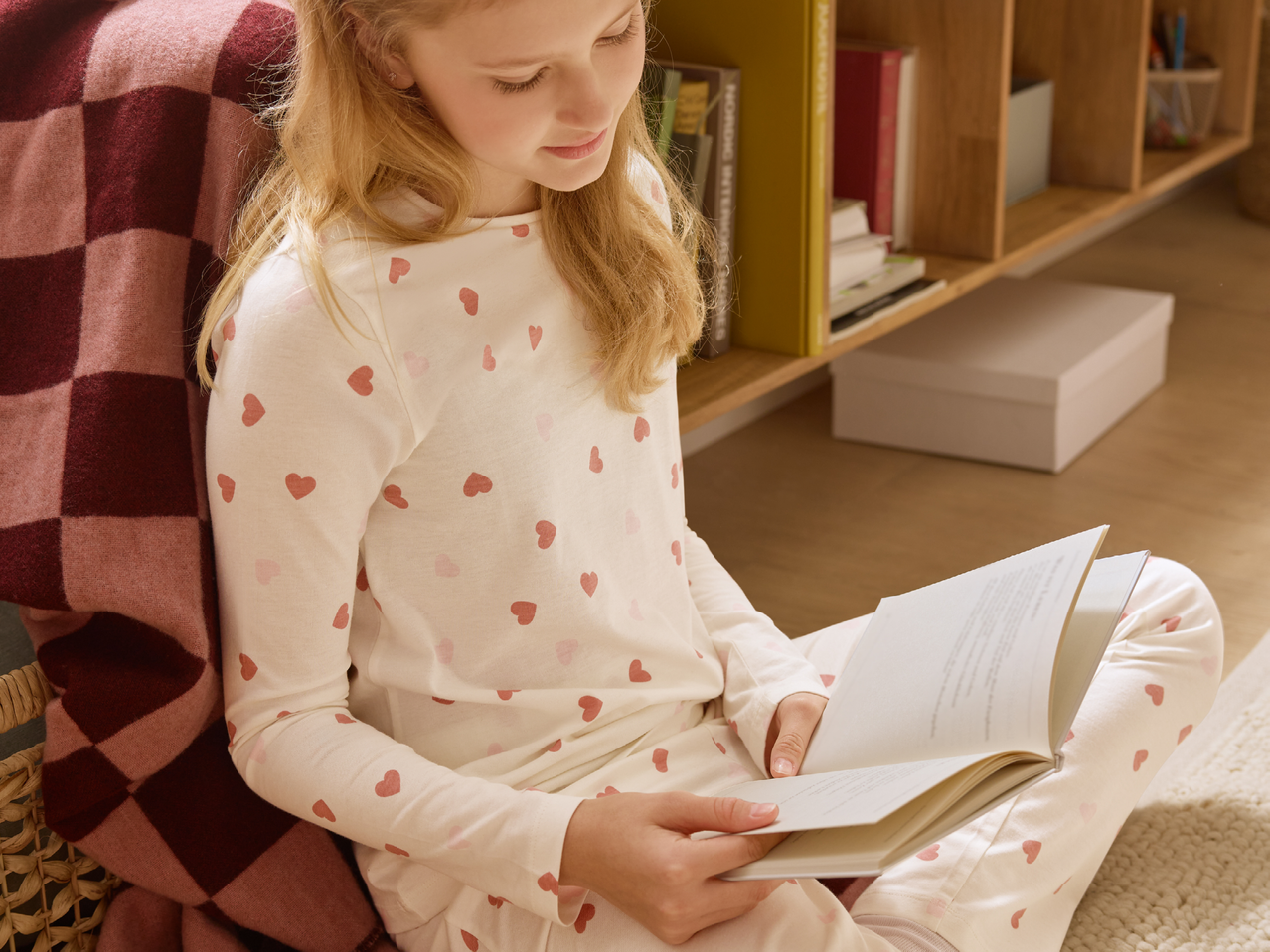Момиче в пижама на сърца чете книга, седнало до карирано одеяло.