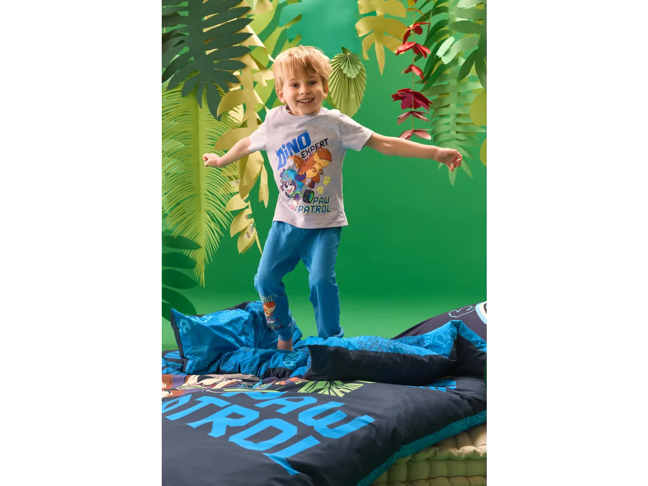 Menino de t-shirt cinzenta e calças de pijama azuis com personagens de desenhos animados, a saltar sobre roupa de cama azul.