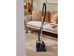 Vacuum cleaner on a textured rug in a living room with a sofa and wooden shelf.