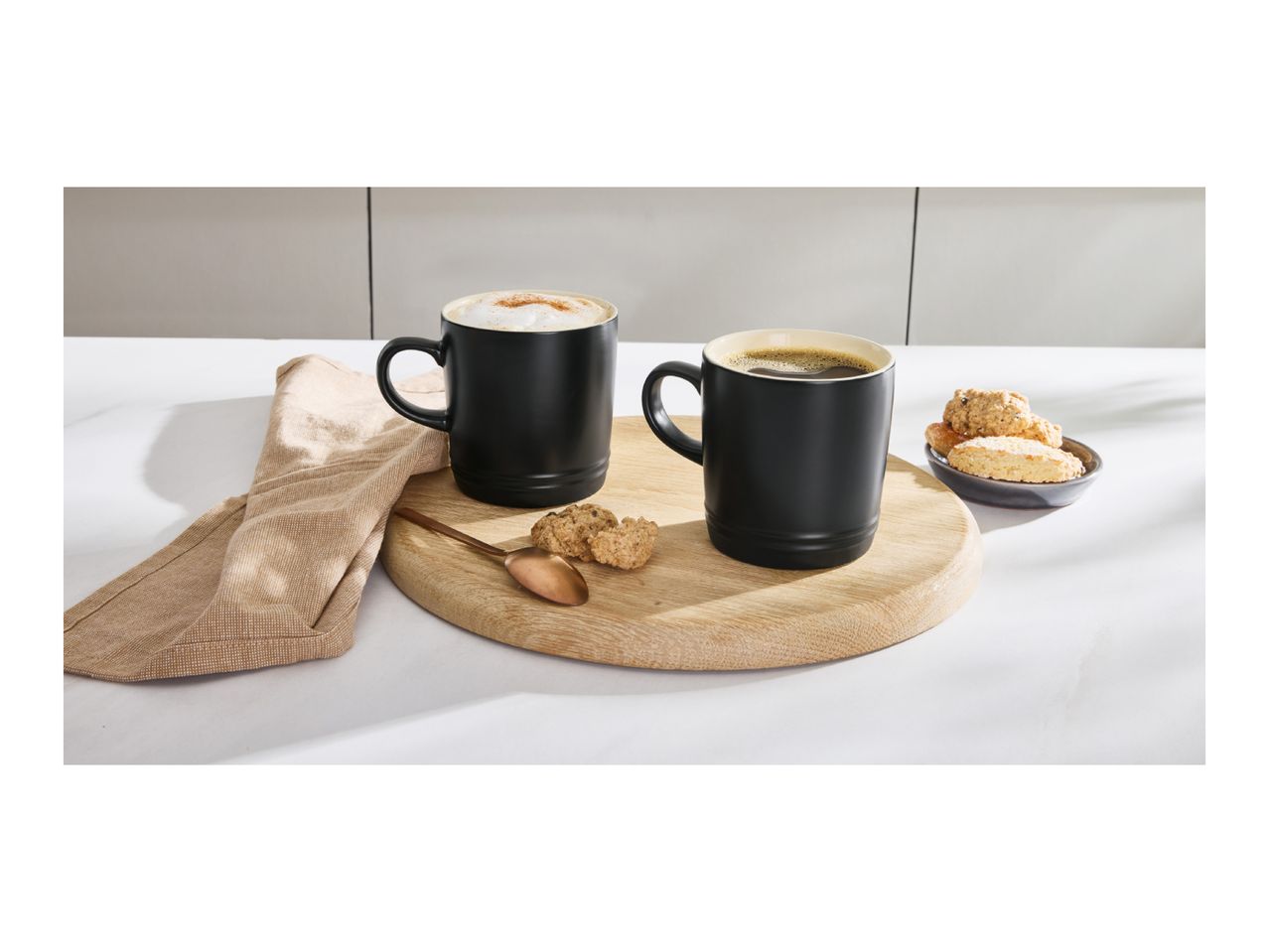 Two black mugs with coffee, one with foam and cinnamon, on a wooden board with cookies.
