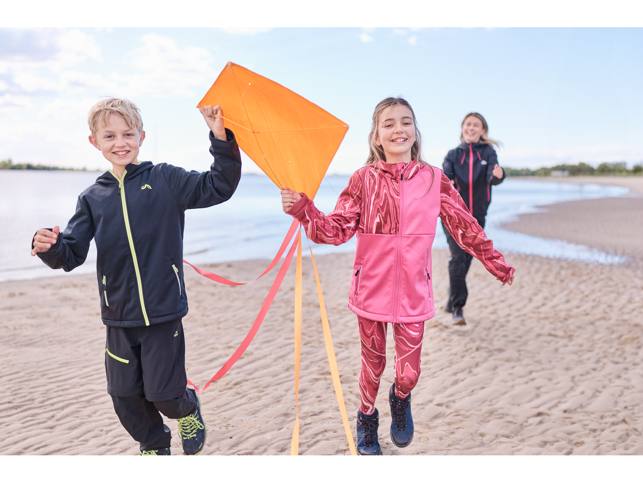 Twee kinderen in Lupilu softshell jassen rennen op het strand met een vlieger, een ander kind volgt.