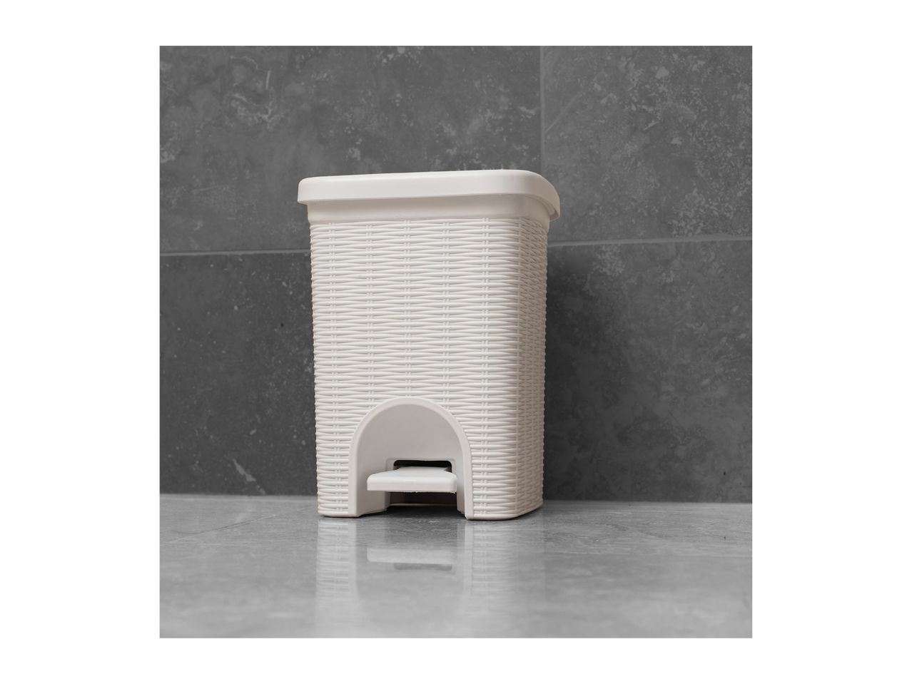 White rattan-style pedal bin on a grey tiled floor against a grey tiled wall.
