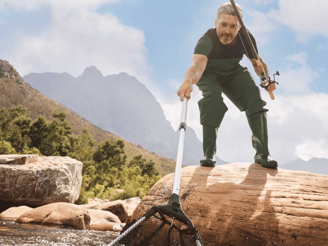 Mann in Wathose und Gummistiefeln mit Angelrute und Kescher beim Angeln in einem Fluss.