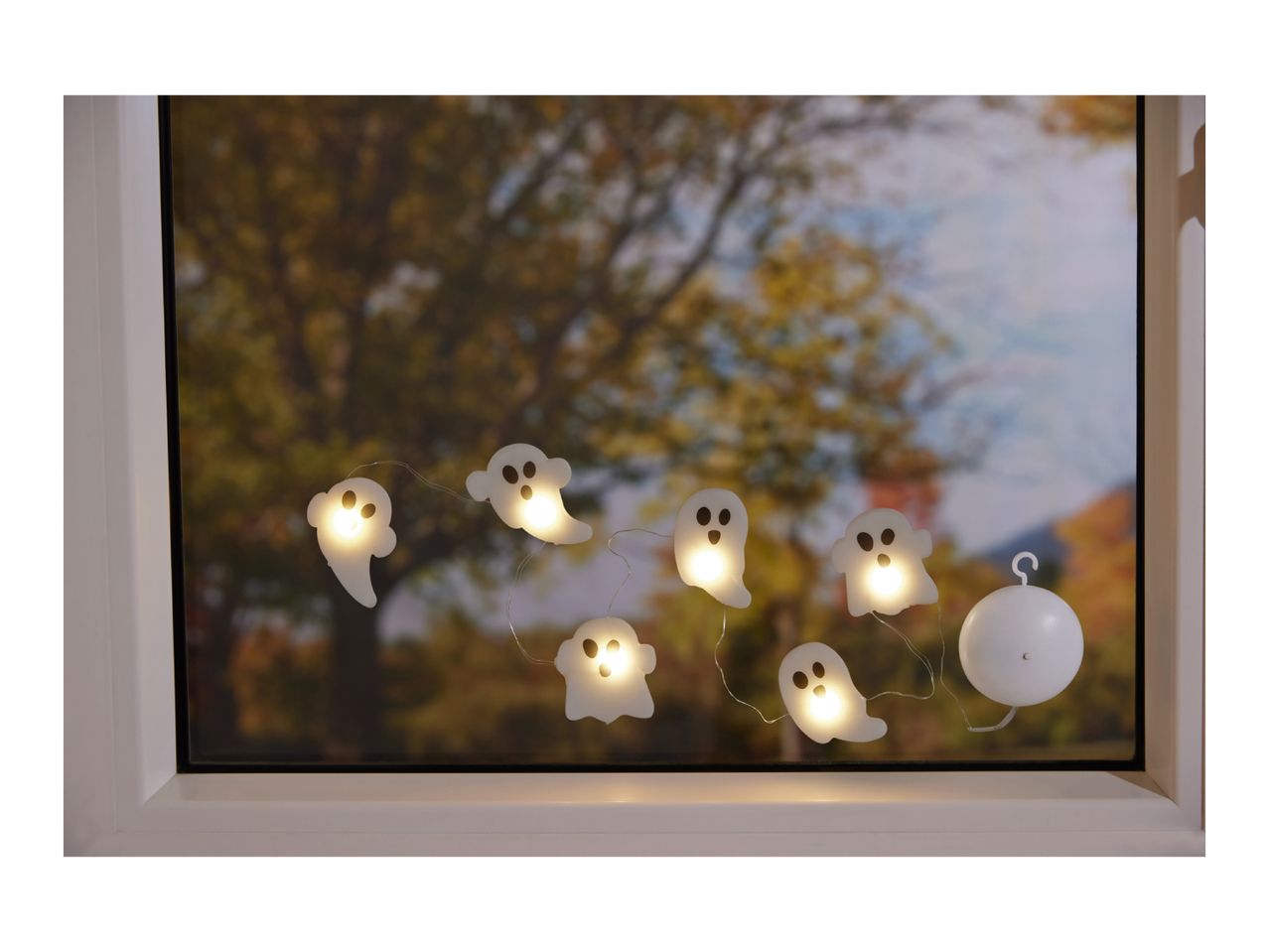 Halloween ghost string lights hanging in a window with autumn trees outside.