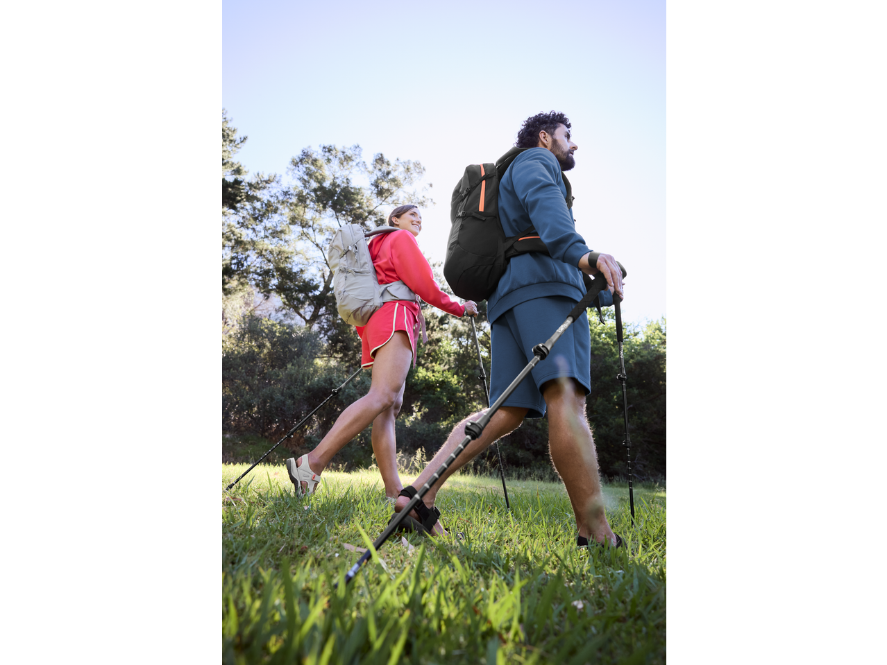 Paar mit Rucksäcken und Trekkingstöcken geht auf Gras.