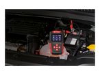 A red and black battery tester, branded Ultimate Speed, connected to a car battery in an engine bay.