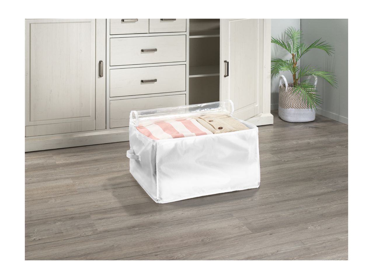 White fabric storage bin with a clear top, filled with clothes, on a wooden floor in front of a white cabinet.