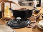 A black cast iron Casserole Dish with meat and sausage, surrounded by kitchenware on a wooden table.