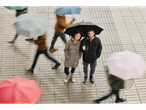 A couple in rain gear stands on a wet sidewalk while blurred people walk by with umbrellas.