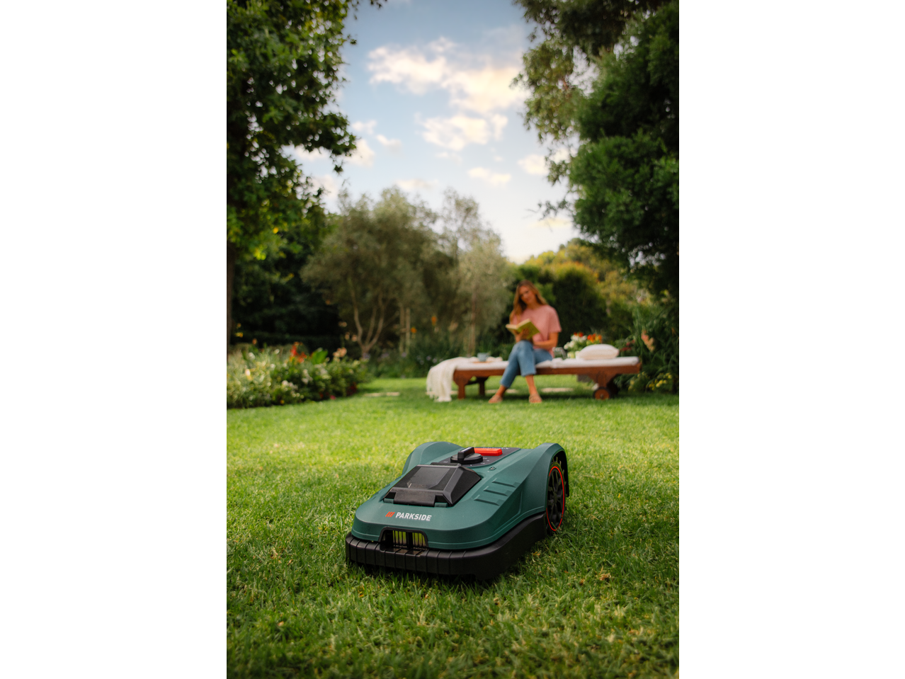 PARKSIDE® 20V Robot Lawn Mower on grass, with a woman reading in the background.