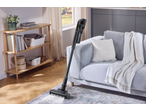 Kabelloser Staubsauger auf einem Teppich in einem Wohnzimmer mit Sofa und Holzregal.