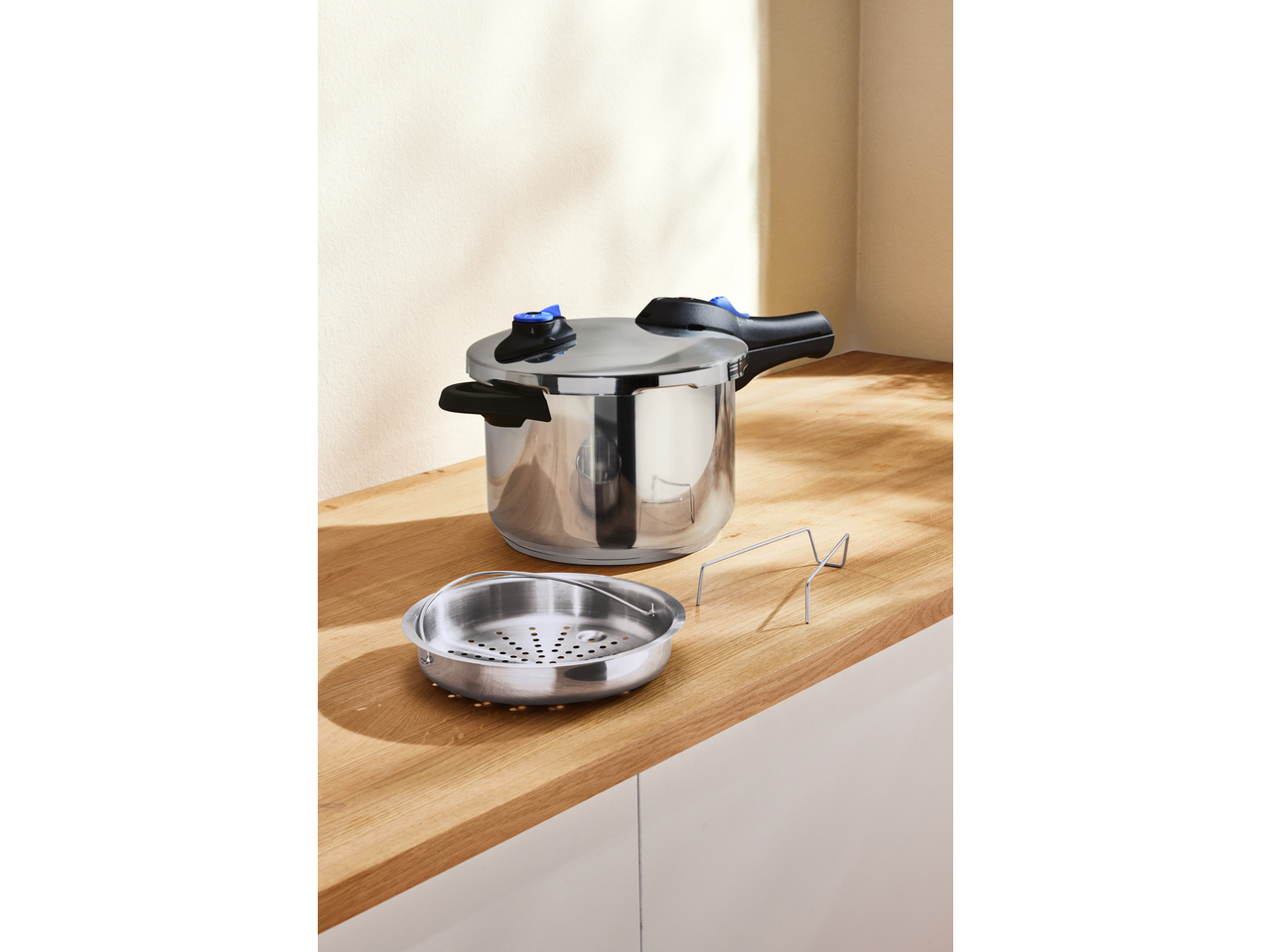 A stainless steel pressure cooker with a blue and black handle, next to a steamer basket.