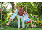 Father and two children playing on swings in a sunny garden with flowers.
