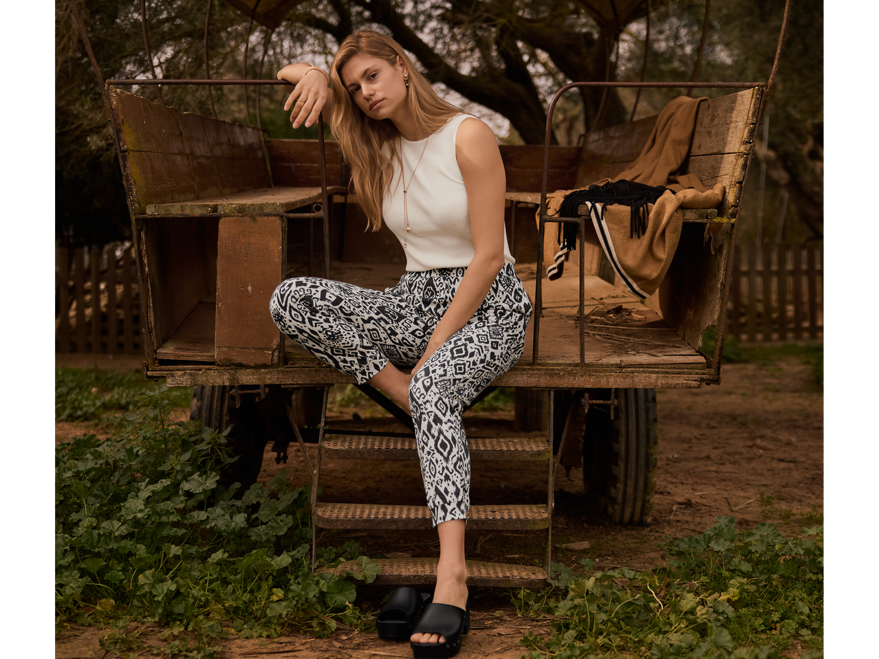 Žena modelira šarene pantalone, beli top i crne papuče na rustikalnom vozu.