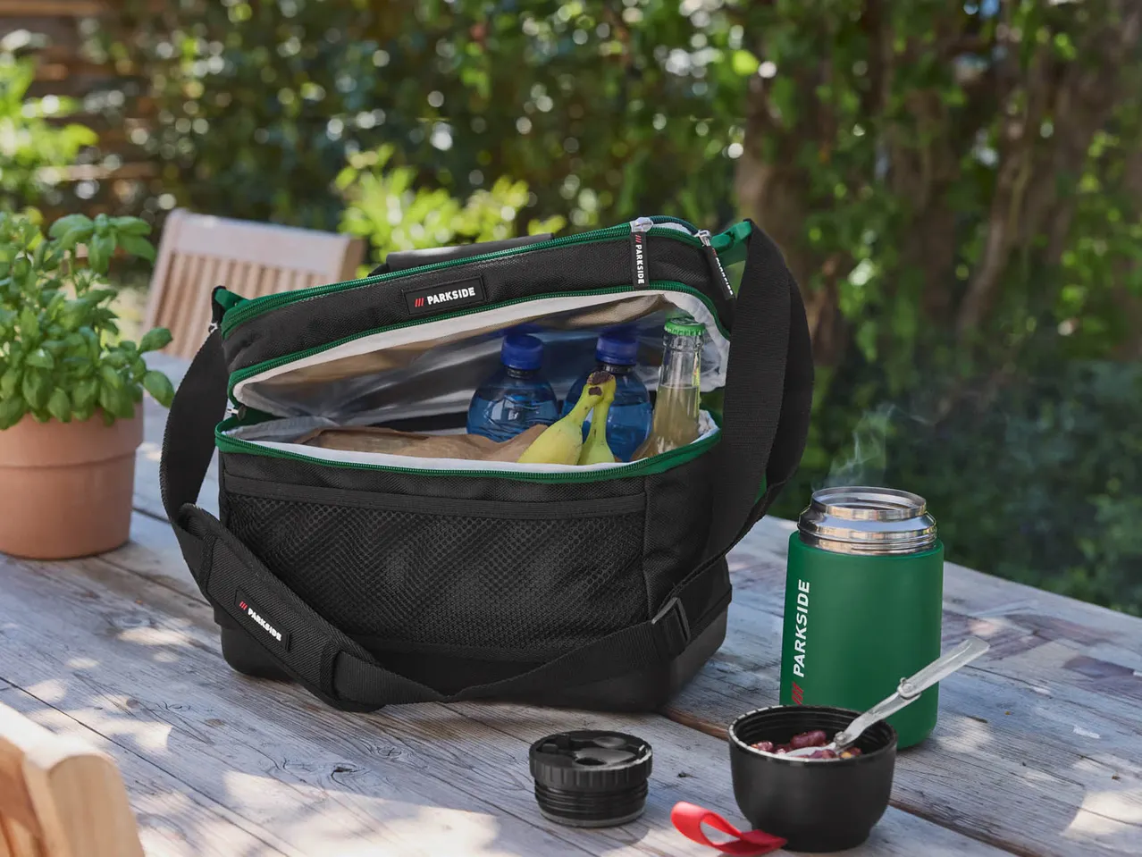 Una borsa frigo Parkside aperta con banane e bevande, accanto a un thermos e una ciotola su un tavolo di legno.