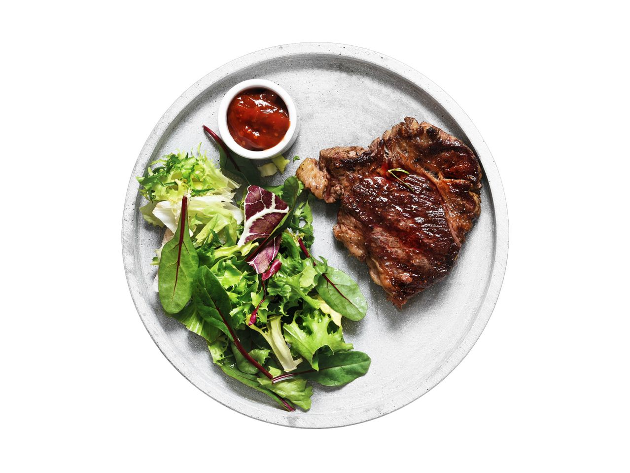 Grilled steak with a side salad and a small bowl of sauce on a grey plate.