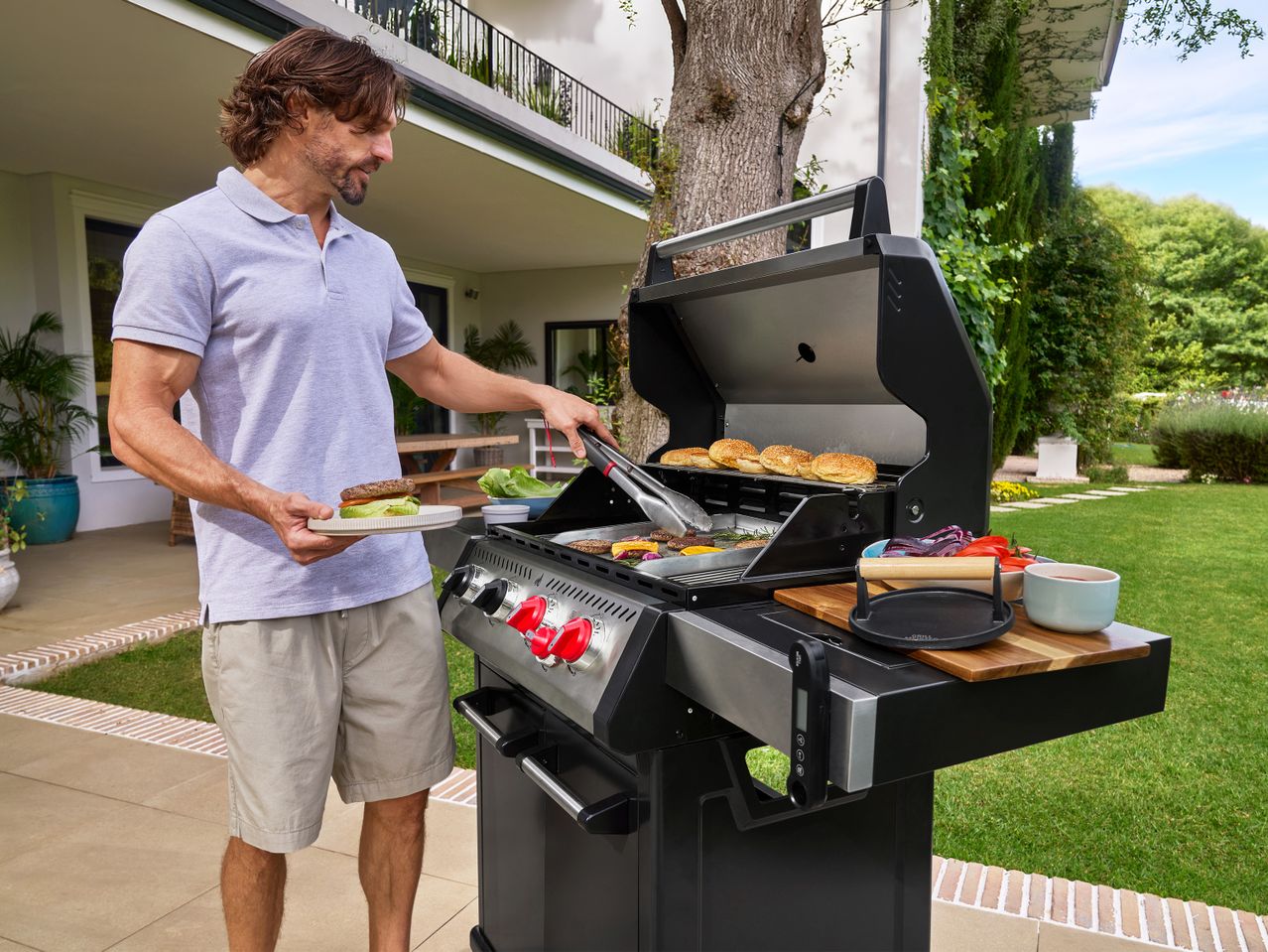 Άνδρας ψήνει μπιφτέκια και ψωμάκια σε ψησταριά υγραερίου σε κήπο.