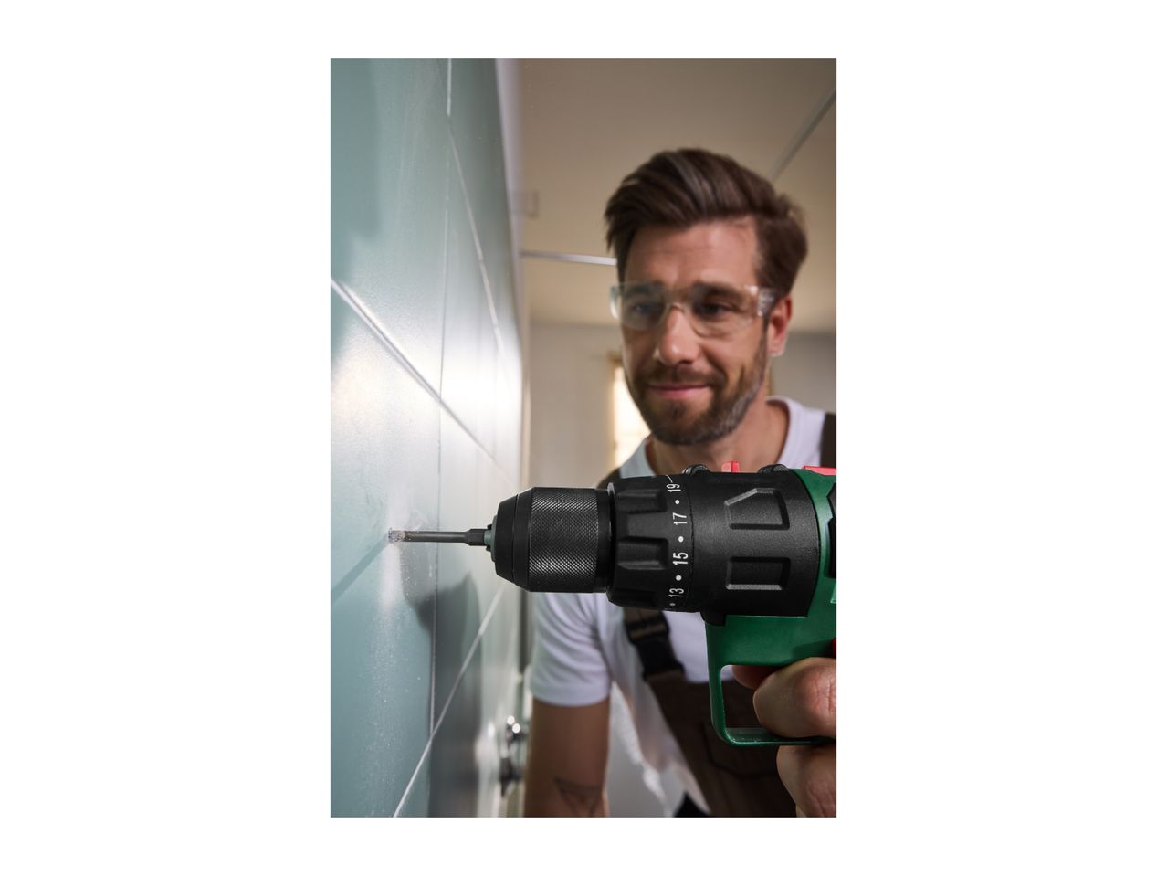 Man drilling into a tiled wall with a cordless drill, wearing safety glasses.