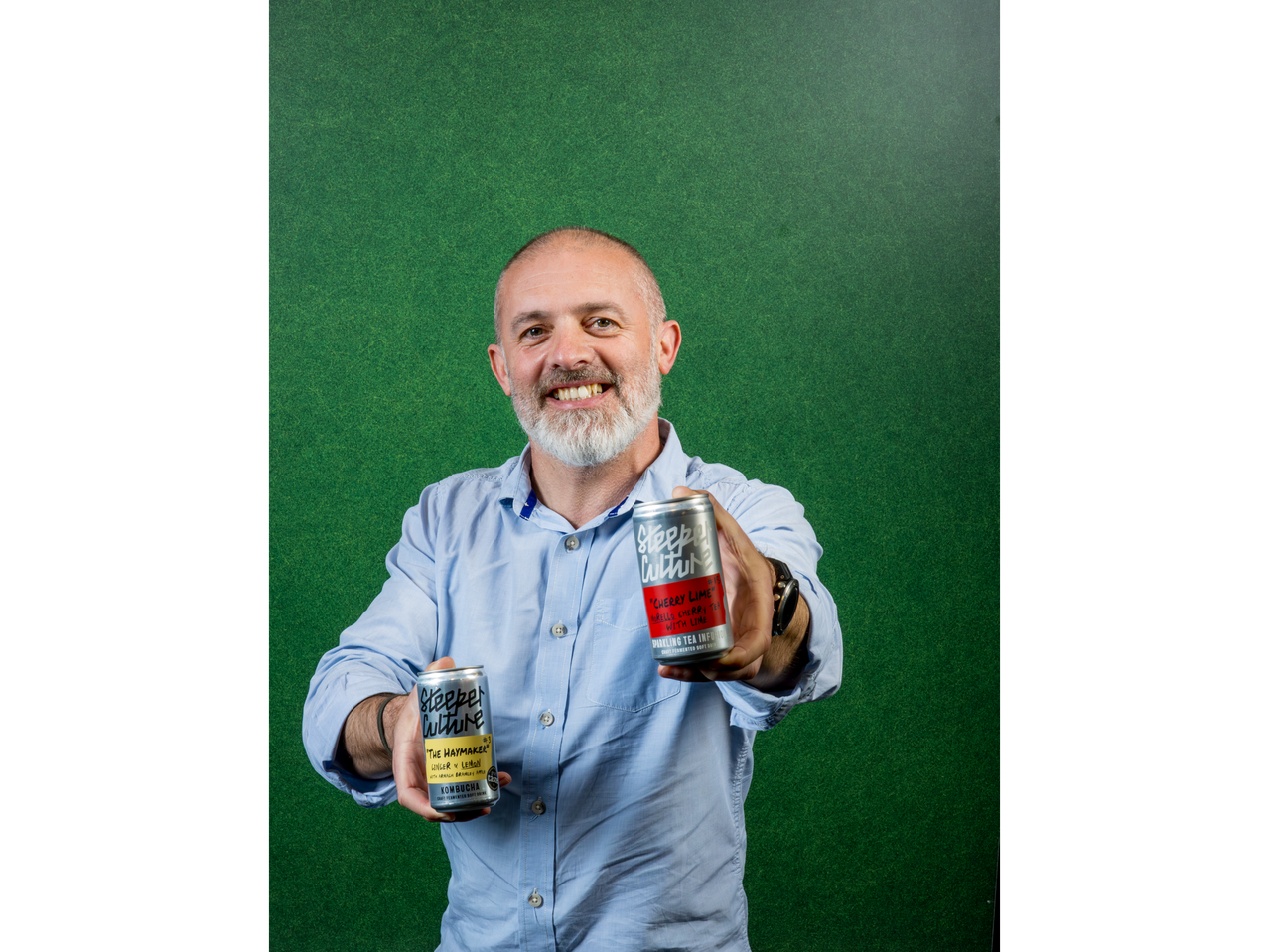 A man holding two Steeper Culture cans, including 'The Haymaker' Ginger & Lemon Kombucha.