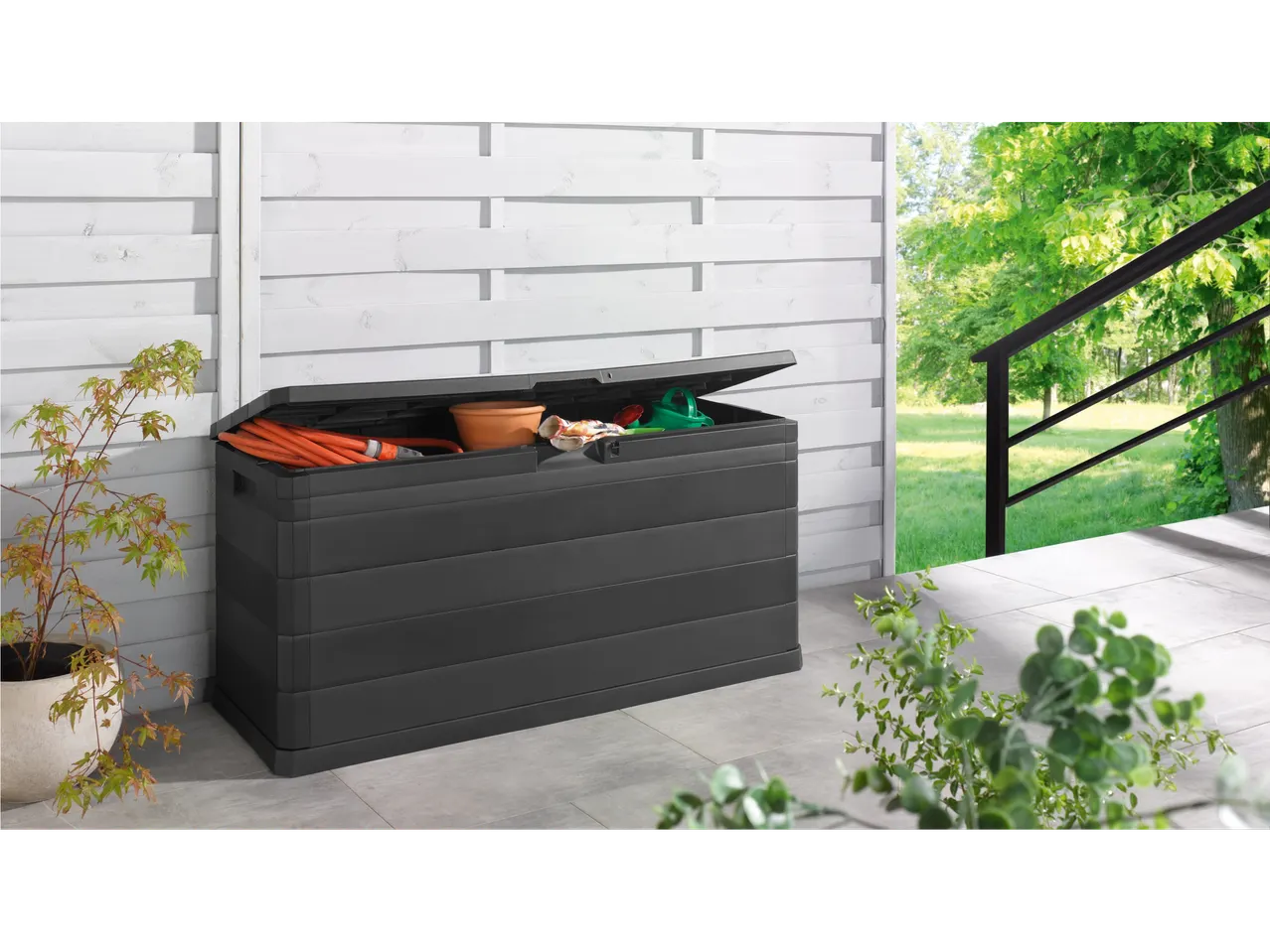 A black outdoor storage box with gardening tools and a hose inside, on a patio.