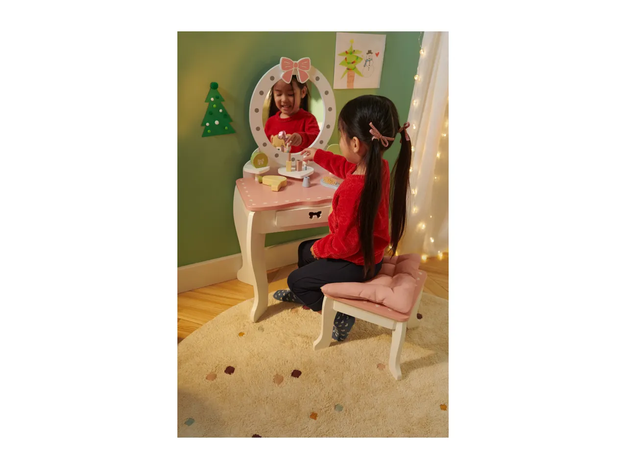 A young girl playing with a pink wooden toy dressing table and stool, decorated for Christmas.