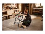 Man measuring wood on a workbench in a workshop, surrounded by tools and wood.