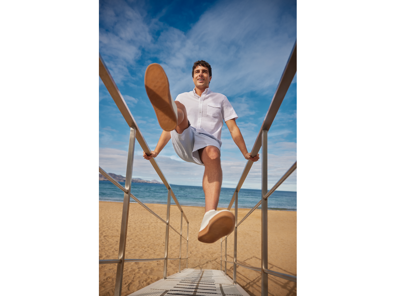 Man in a white seersucker shirt and shorts, stepping down beach stairs.