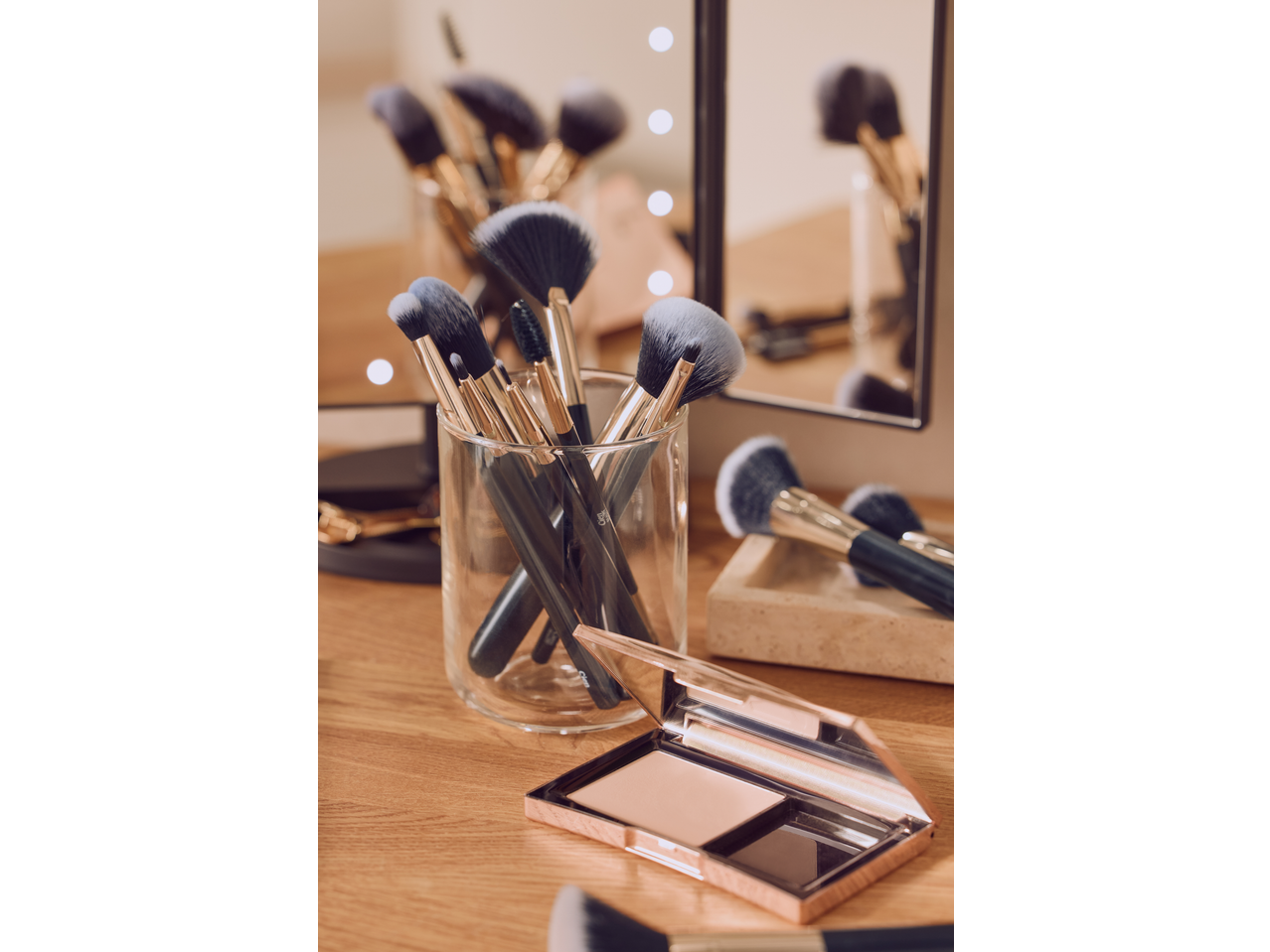 Makeup brushes in a glass holder and an open compact powder on a wooden surface.