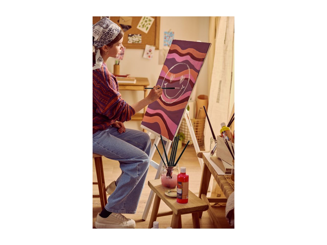 Young woman painting a smiley face on canvas, surrounded by art supplies.