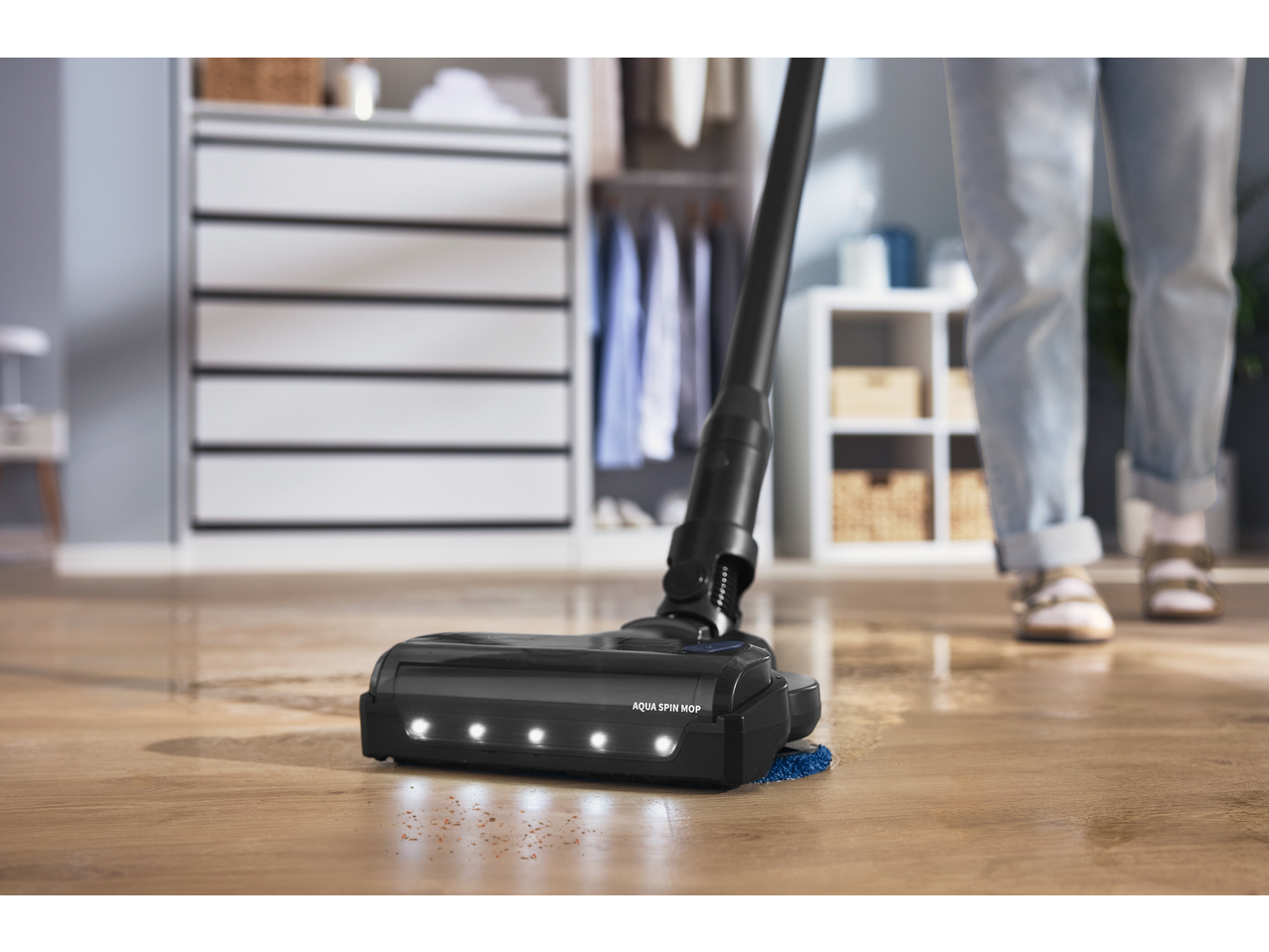 Cordless vacuum cleaner with Aqua Spin Mop head cleaning a wooden floor with visible dirt.