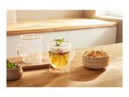 Two double-walled glass mugs, one with tea and mint, next to a bowl of cookies on a wooden surface.