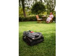Robotic lawnmower on green grass with a woman relaxing in the background.