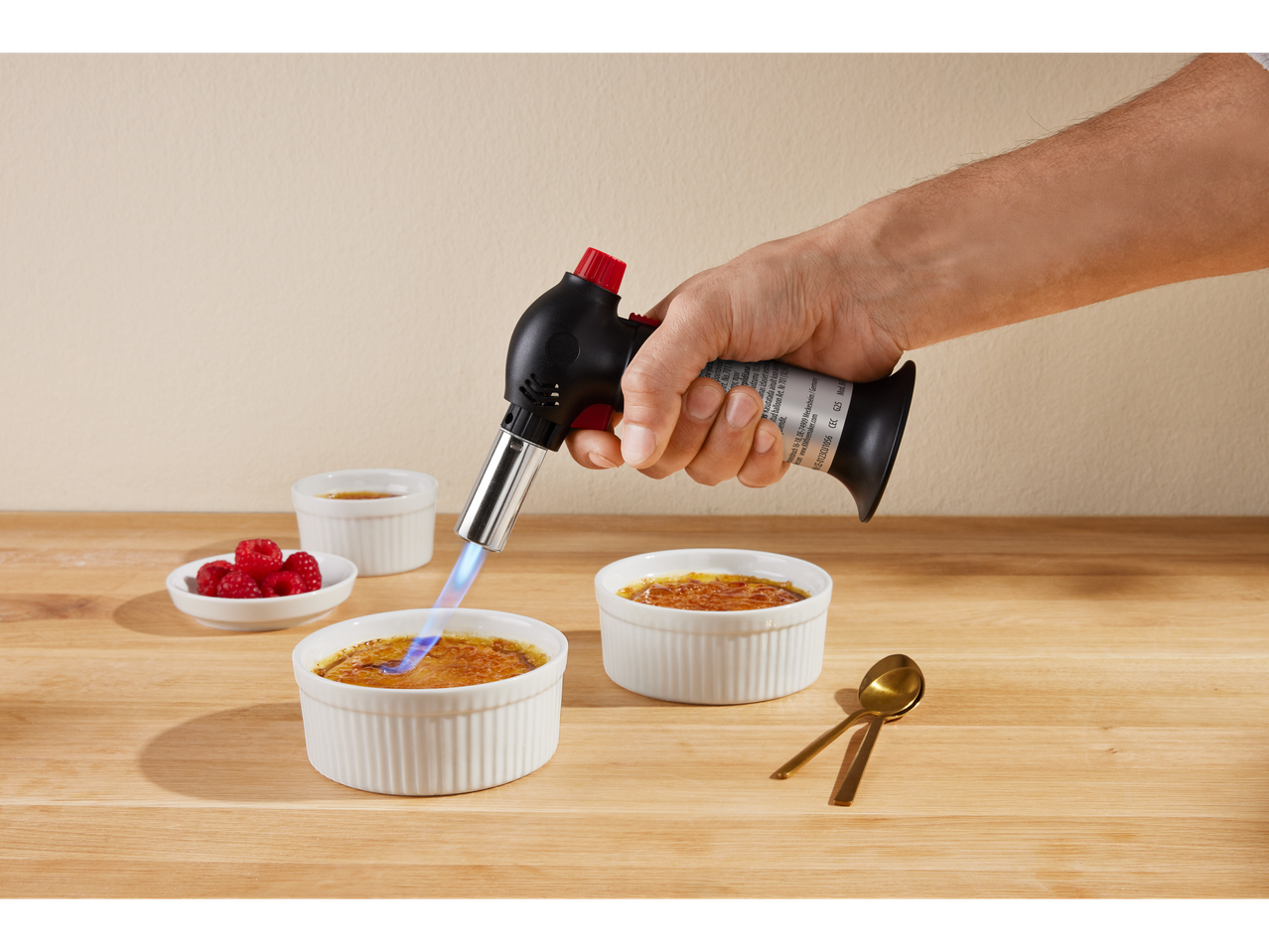 Käsi põletab köögipõletiga crème brûlée'd, vaarikate ja lusikatega puidust pinnal.