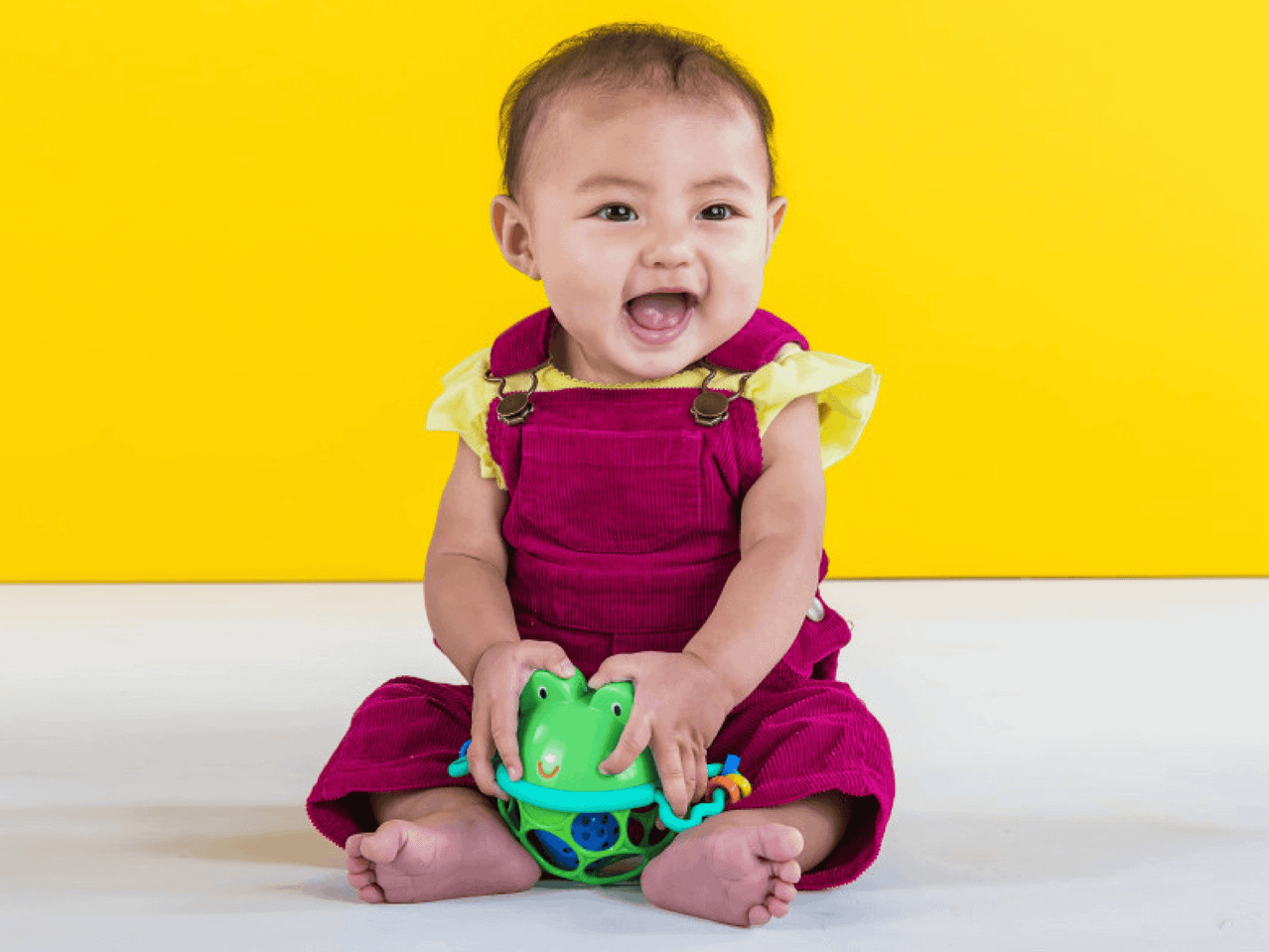 Ein glückliches Baby in weinroten Stramplern spielt mit einem grünen Spielzeug.