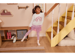 Djevojčica u ružičastoj haljini i tajicama smiješi se pored stepenica u dječjoj sobi.